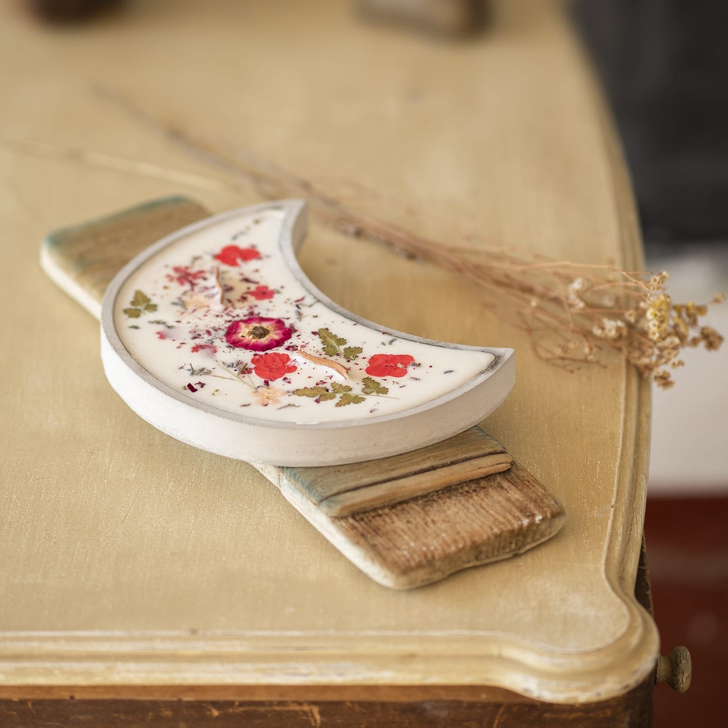 La Vela Aromática Fleur-de-Bois en Cuenco de Cemento en Forma de LUNA con Mecha Ondulada, decorada con flores y hierbas secas, descansa sobre un pequeño cepillo de madera en una mesa beige vintage junto a un manojo de ramas secas.