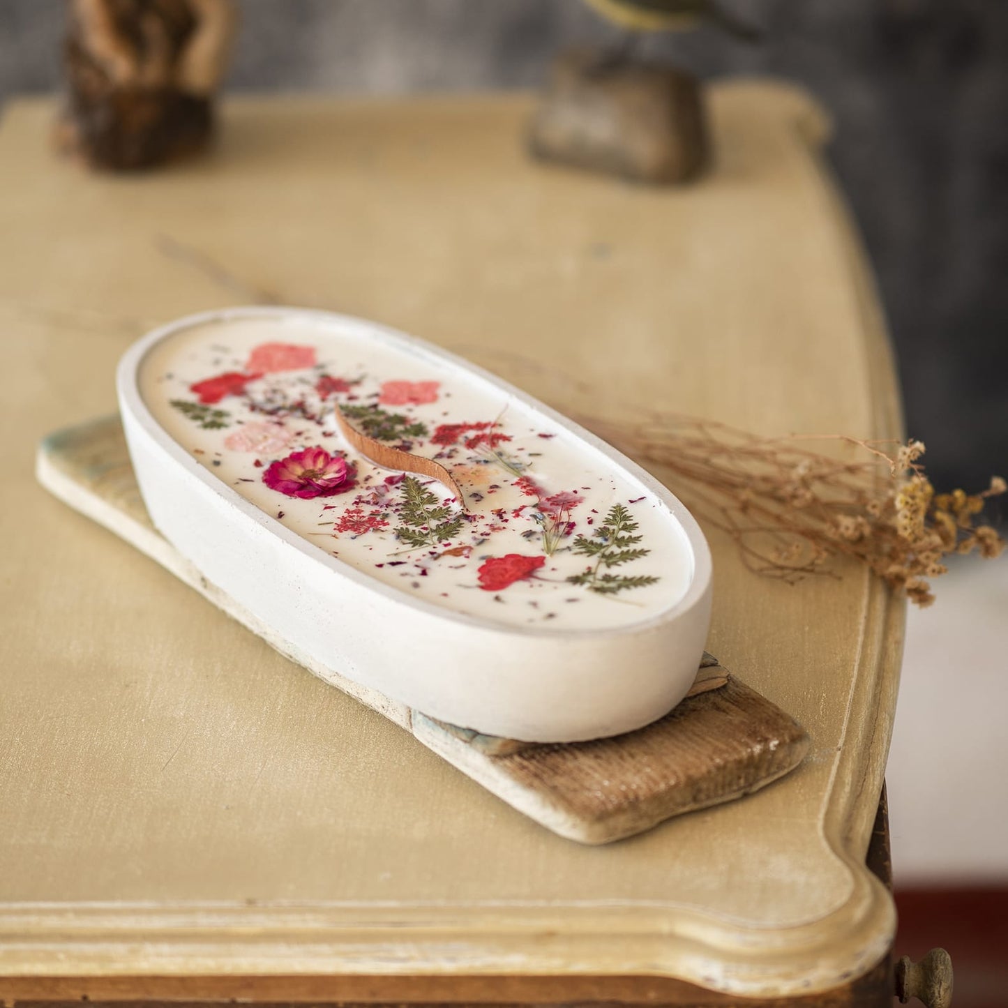 La Vela Aromatica en Cuenco de Cemento Ovalado GRANDE con Mecha Ondulada de Fleur-de-Bois, decorada con flores y hojas secas, reposa sobre una tabla de madera en una mesa vintage; al fondo se ven difusas figuras decorativas.