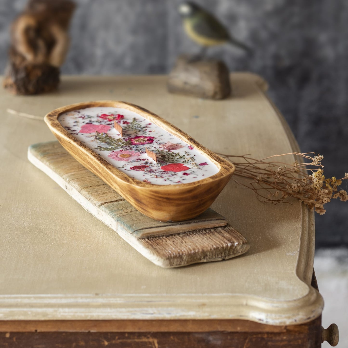 La Vela Aromática en Cuenco de Madera Grande con Mecha Ondulada de Fleur-de-Bois, decorada con flores y hierbas secas en cera de soja vegana, reposa sobre una mesa rústica junto a una tabla de cortar, acompañada por un ave decorativa y una planta seca.