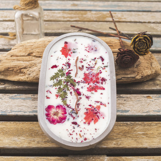 La vela aromática en cuenco de cemento ovalado con mecha ondulada de Fleur-de-Bois reposa sobre madera junto a vidrio y rosas secas, siendo un detalle especial e ideal para regalar.
