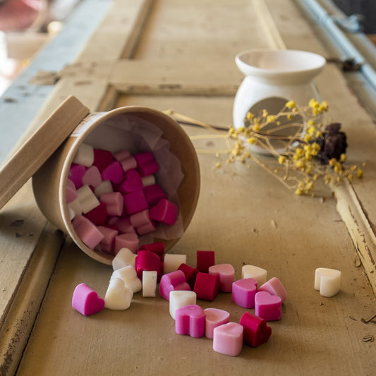 Un recipiente de cera fundida Fleur-de-Bois Esencias en Miniatura HEART WISHES derrama cera fundida roja, rosa y blanca en forma de corazón sobre una superficie de madera, con un soporte de cerámica blanca y flores de primavera amarillas secas cerca.