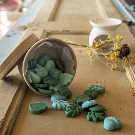 Un vaso con punta derrama sobre madera rústica las Esencias de Cera en Miniatura HOJAS SUELTAS de Fleur-de-Bois. Un calentador de cera de cerámica y flores secas sugieren las fragancias artesanales de Fleur-de-Bois.