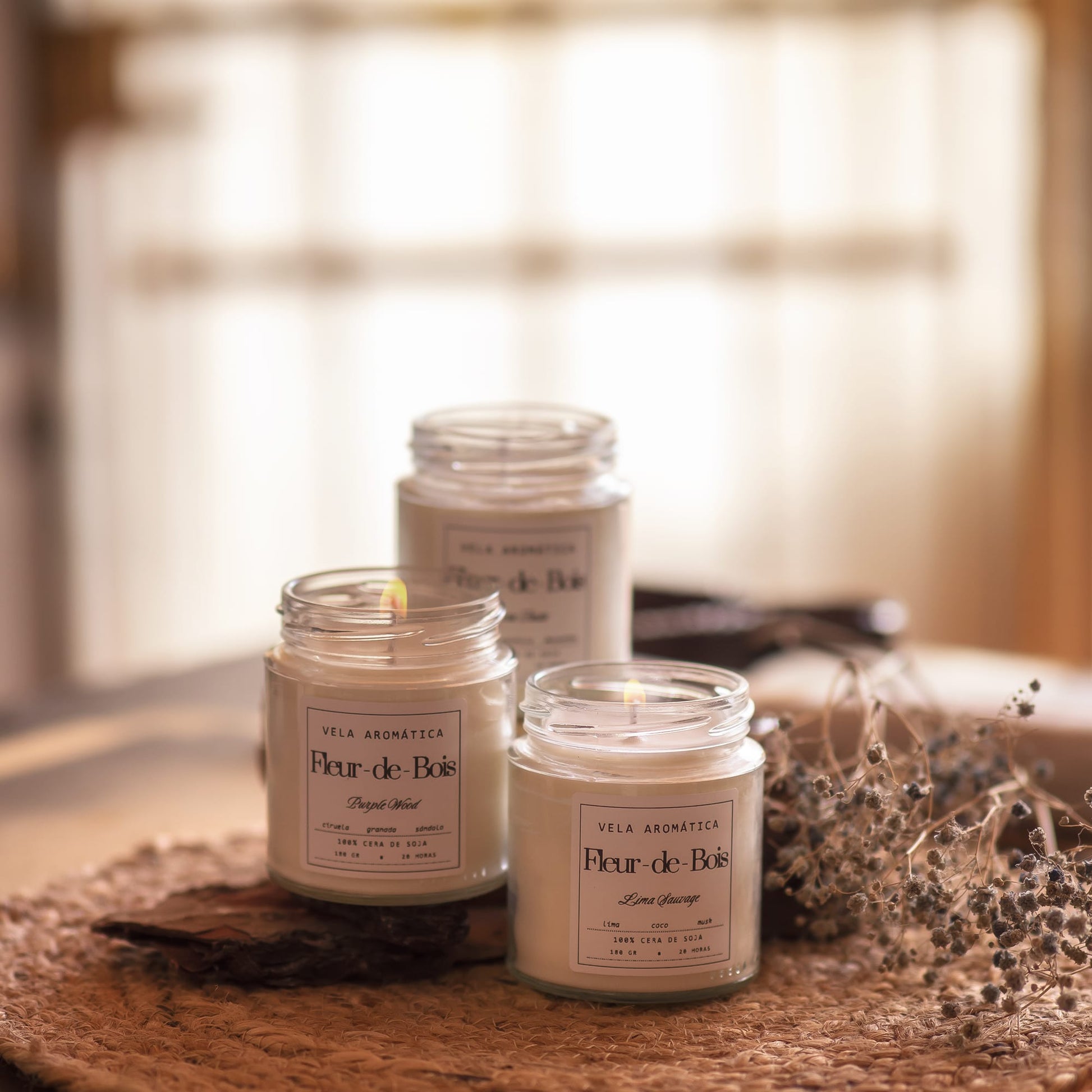 Tres velas encendidas Fleur-de-Bois Vela Aromática Signature (180 g) se posan sobre una estera tejida con flores secas, añadiendo una acogedora decoración de hogar y calidez a la escena.