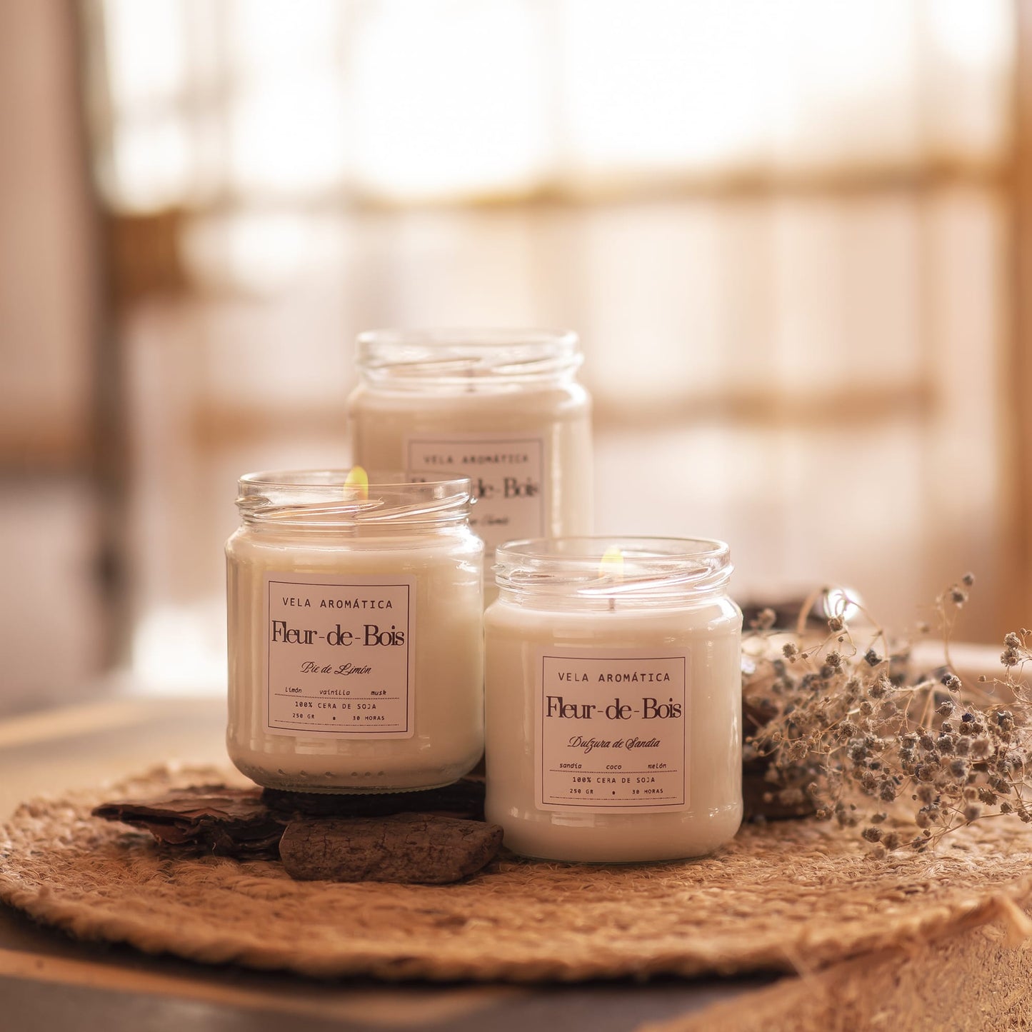 Tres velas de soja Fleur-de-Bois Signature Vela Aromática de 250 g encendidas en tarros de cristal se asientan sobre una alfombra tejida con flores secas, sobre un fondo cálido y difuminado que muestra a la perfección la belleza de estas velas hechas a mano.