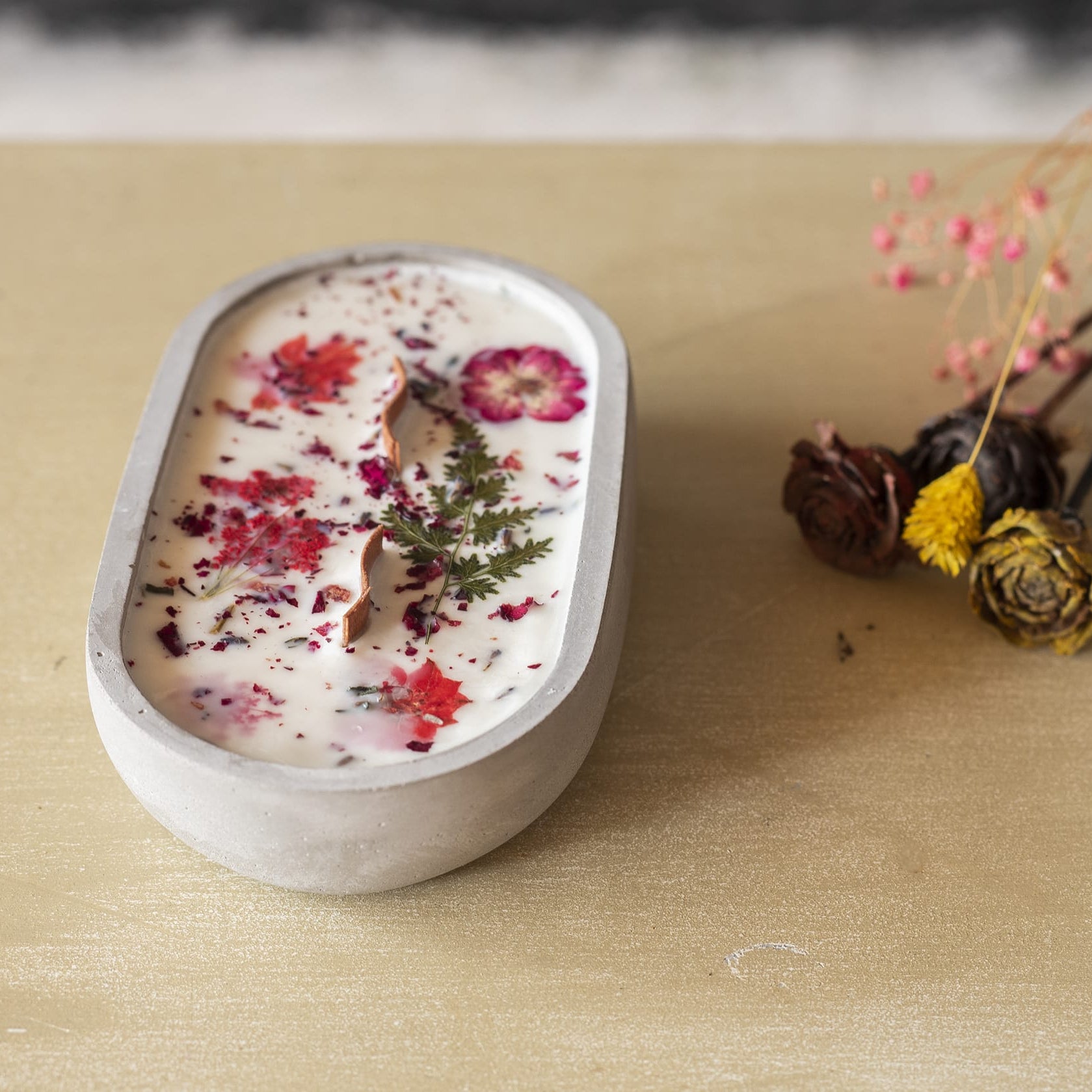 La Vela Aromatica en Cuenco de Cemento Ovalado con Mecha Ondulada de Fleur-de-Bois, decorada con flores y hojas secas, descansa sobre madera clara, acompañada de un ramo seco-ideal como regalo especial.