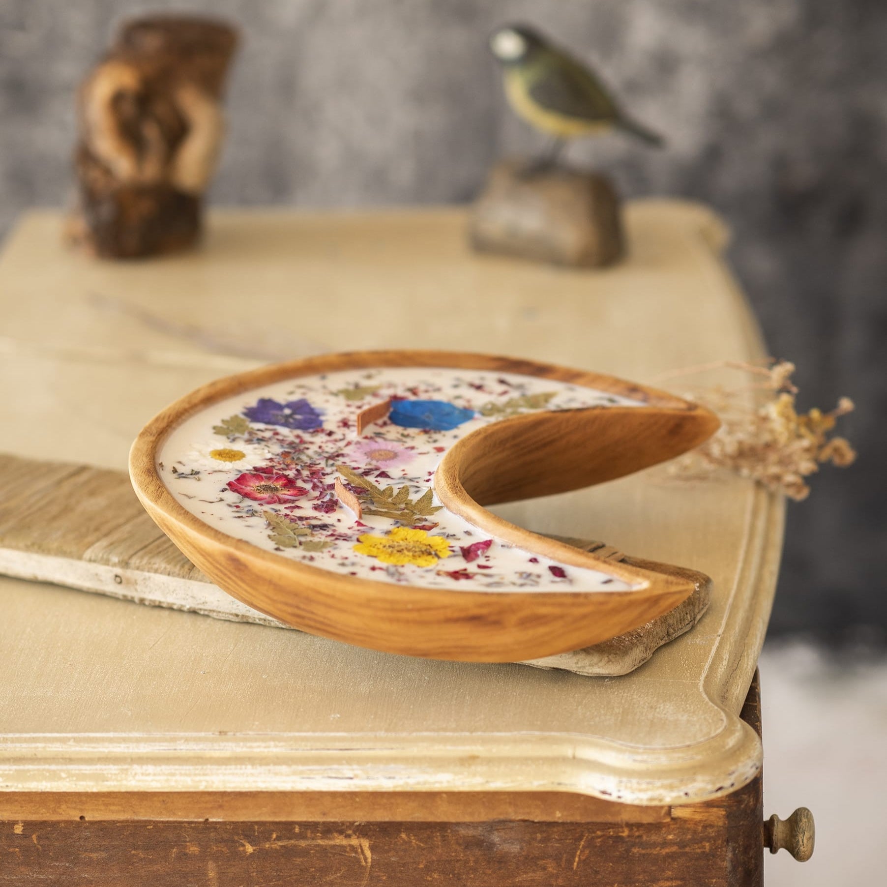 Una Vela Aromática Fleur-de-Bois en Cuenco de Madera en Forma de LUNA con Mecha Ondulada, adornada con flores secas y hierbas, se encuentra en una mesa vintage junto a una figura de pájaro y decoración de madera.
