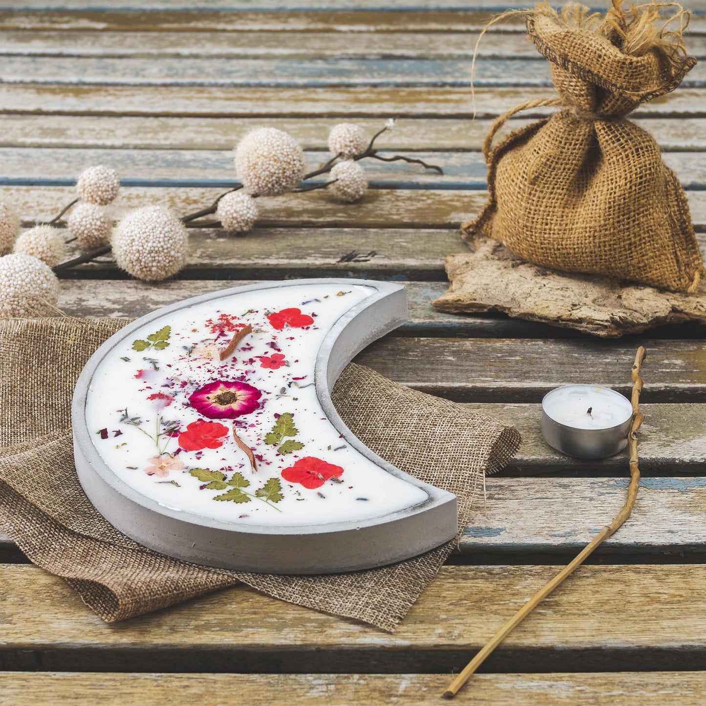 La Vela Aromática Fleur-de-Bois en Cuenco de Cemento en Forma de LUNA con Mecha Ondulada, decorada con flores secas, descansa sobre madera junto a yute, un saquito, ramas de bayas blancas, una vela de té y un incienso-un regalo especial.
