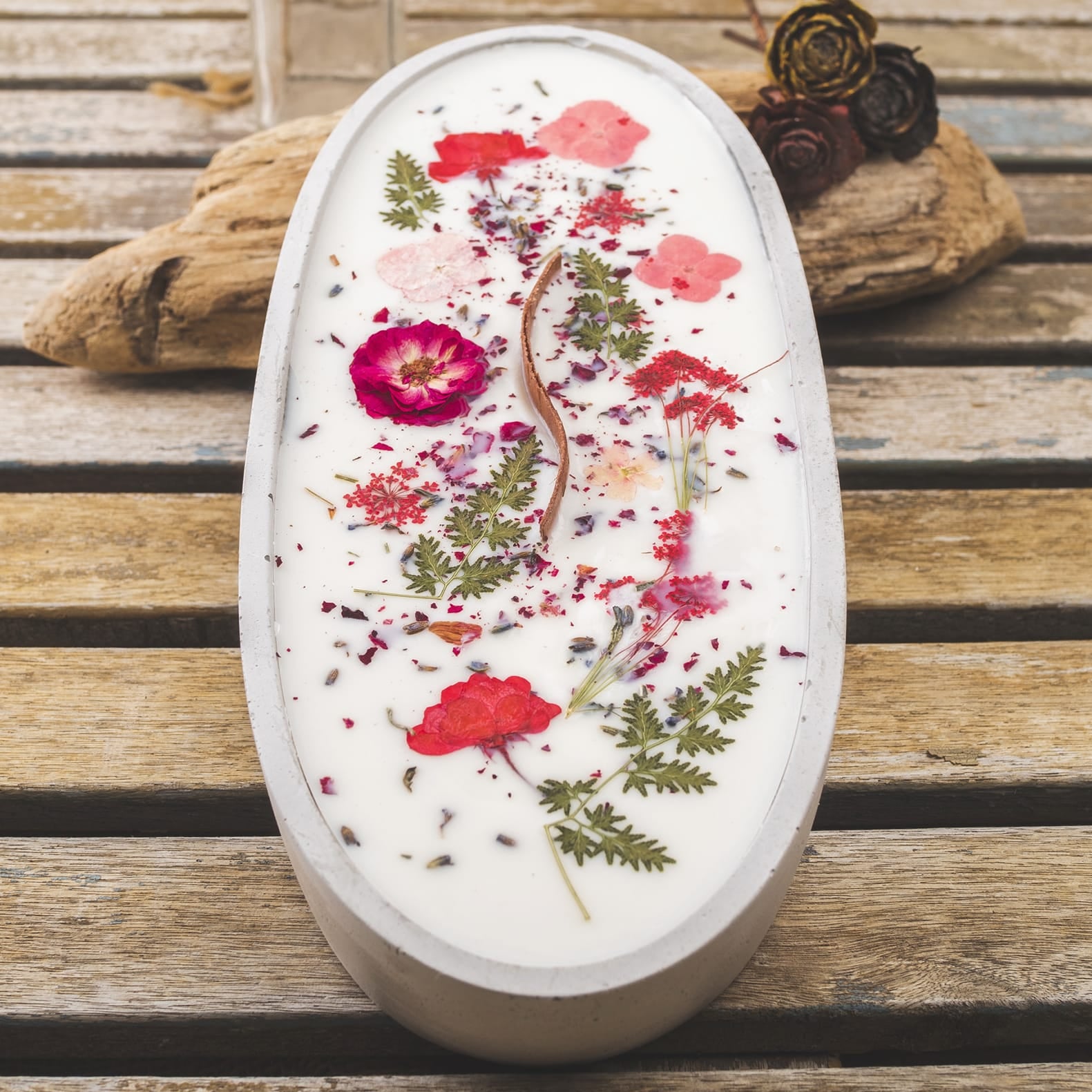 Sobre una mesa de madera descansa la Vela Aromática en Cuenco de Cemento Ovalado GRANDE con Mecha Ondulada de Fleur-de-Bois, decorada con flores secas, hojas y pétalos; al fondo, hay una botella de vidrio y piezas de madera flotante.