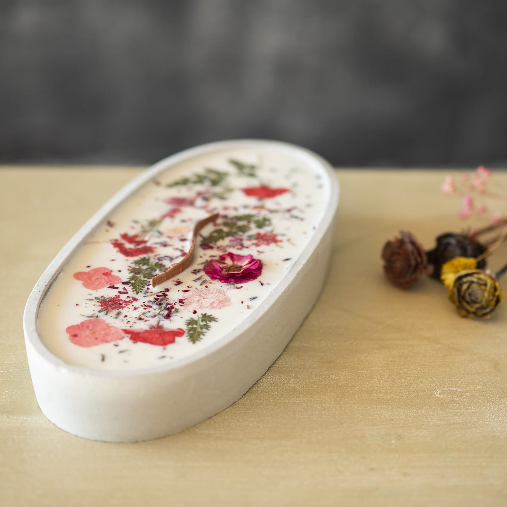 La Vela Aromática en Cuenco de Cemento Ovalado GRANDE de Fleur-de-Bois, con mecha ondulada, decorada con flores secas y pétalos rojos, reposa sobre madera clara; al fondo, flores secas desenfocadas.