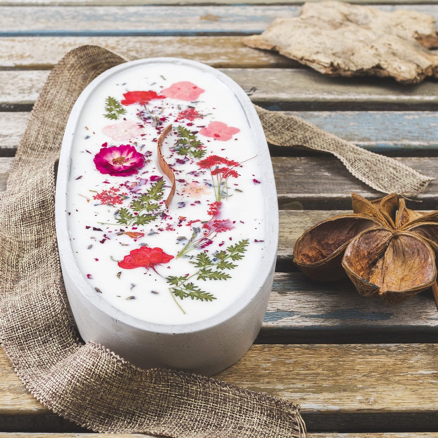 La Vela Aromática Fleur-de-Bois en Cuenco de Cemento Ovalado GRANDE, con mecha ondulada y decorada con flores secas, reposa sobre madera junto a un lazo de yute, una semilla en estrella y un trozo de corteza.