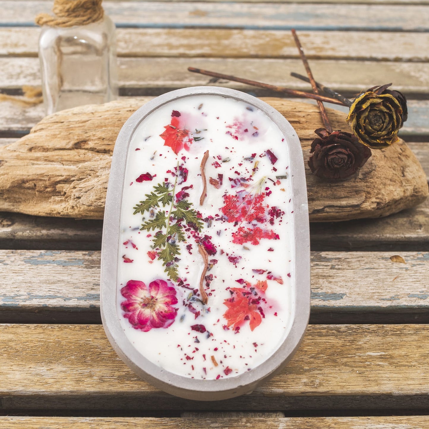 La vela aromática en cuenco de cemento ovalado con mecha ondulada de Fleur-de-Bois reposa sobre madera junto a vidrio y rosas secas, siendo un detalle especial e ideal para regalar.