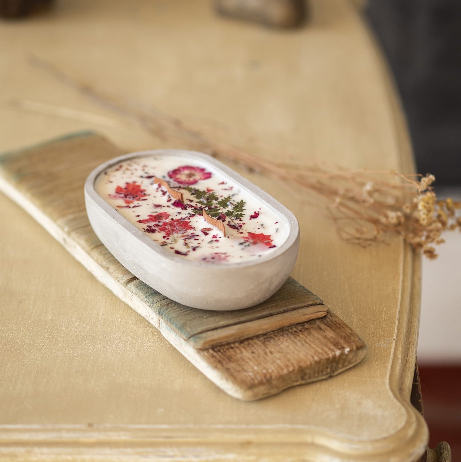 La Vela Aromatica Fleur-de-Bois en un Cuenco de Cemento Ovalado con Mecha Ondulada, adornada con flores secas y hierbas, descansa sobre una bandeja de madera en una mesa beige-ideal como regalo especial.