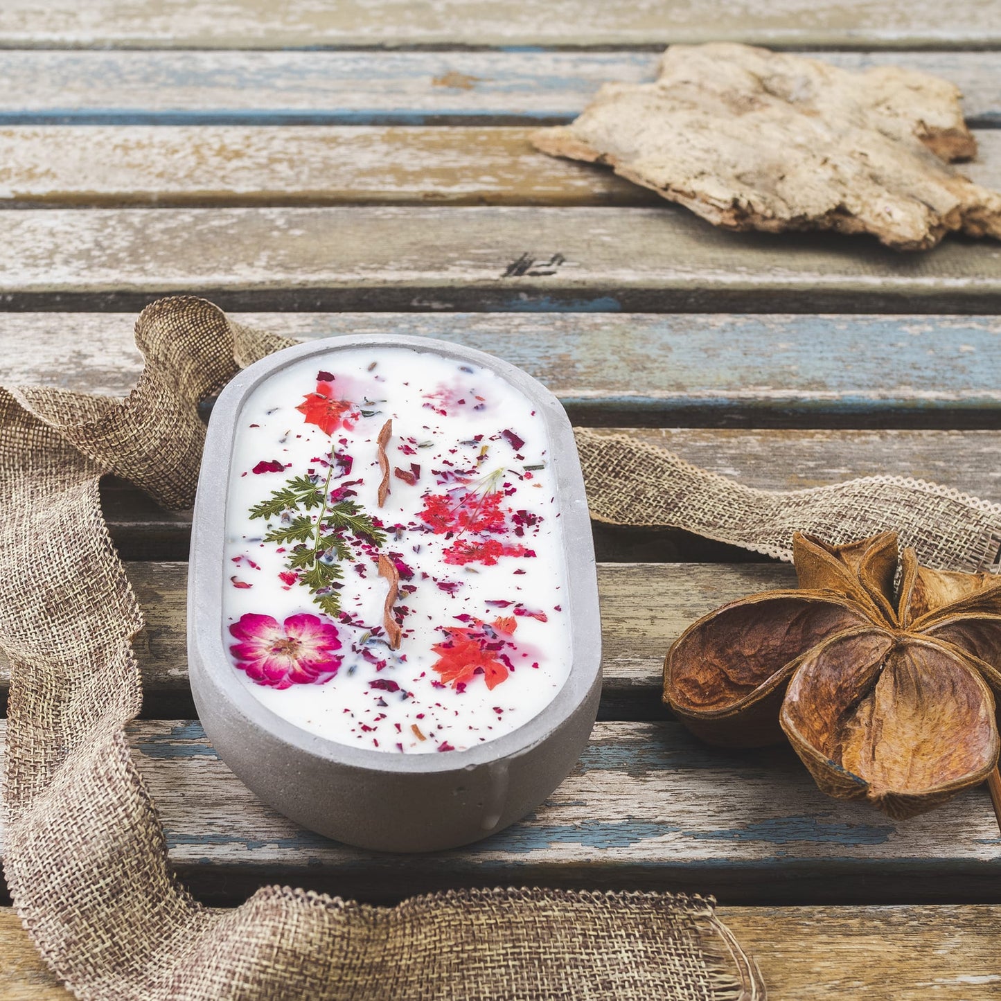 La Vela Aromática Fleur-de-Bois en Cuenco de Cemento Ovalado con Mecha Ondulada, decorada con flores y hojas secas, descansa sobre una mesa rústica-un regalo especial y elegante para cualquier ocasión.