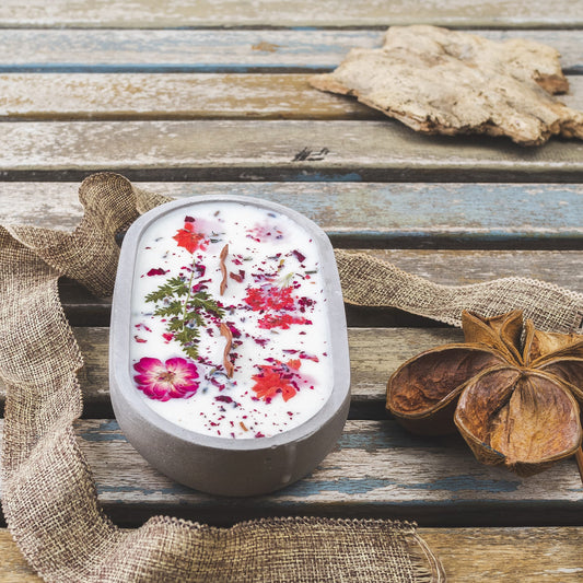 La Vela Aromática Fleur-de-Bois en Cuenco de Cemento Ovalado con Mecha Ondulada, decorada con flores y hojas secas, descansa sobre una mesa rústica-un regalo especial y elegante para cualquier ocasión.