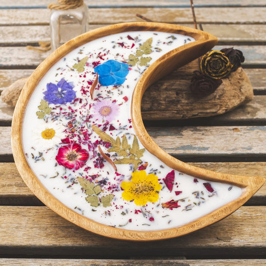 La Vela Aromatica Fleur-de-Bois en Cuenco de Madera en Forma de LUNA con Mecha Ondulada, adornada con flores y hojas secas, descansa sobre madera rústica; botánicos secos y una botella con corcho aparecen al fondo.
