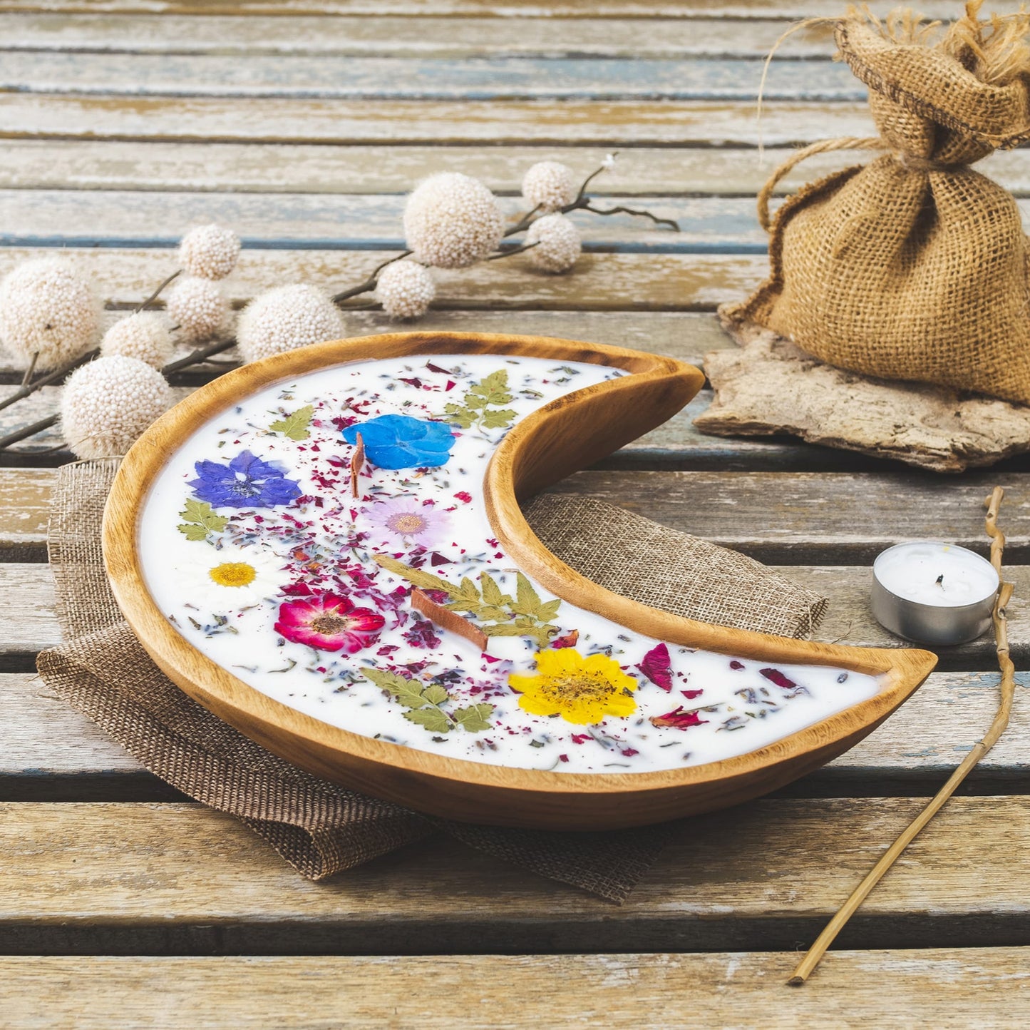 La Fleur-de-Bois Vela Aromatica en Cuenco de Madera en Forma de LUNA con Mecha Ondulada, decorada con flores secas, reposa sobre una mesa rústica junto a ramas, un saco de yute y otros adornos naturales.