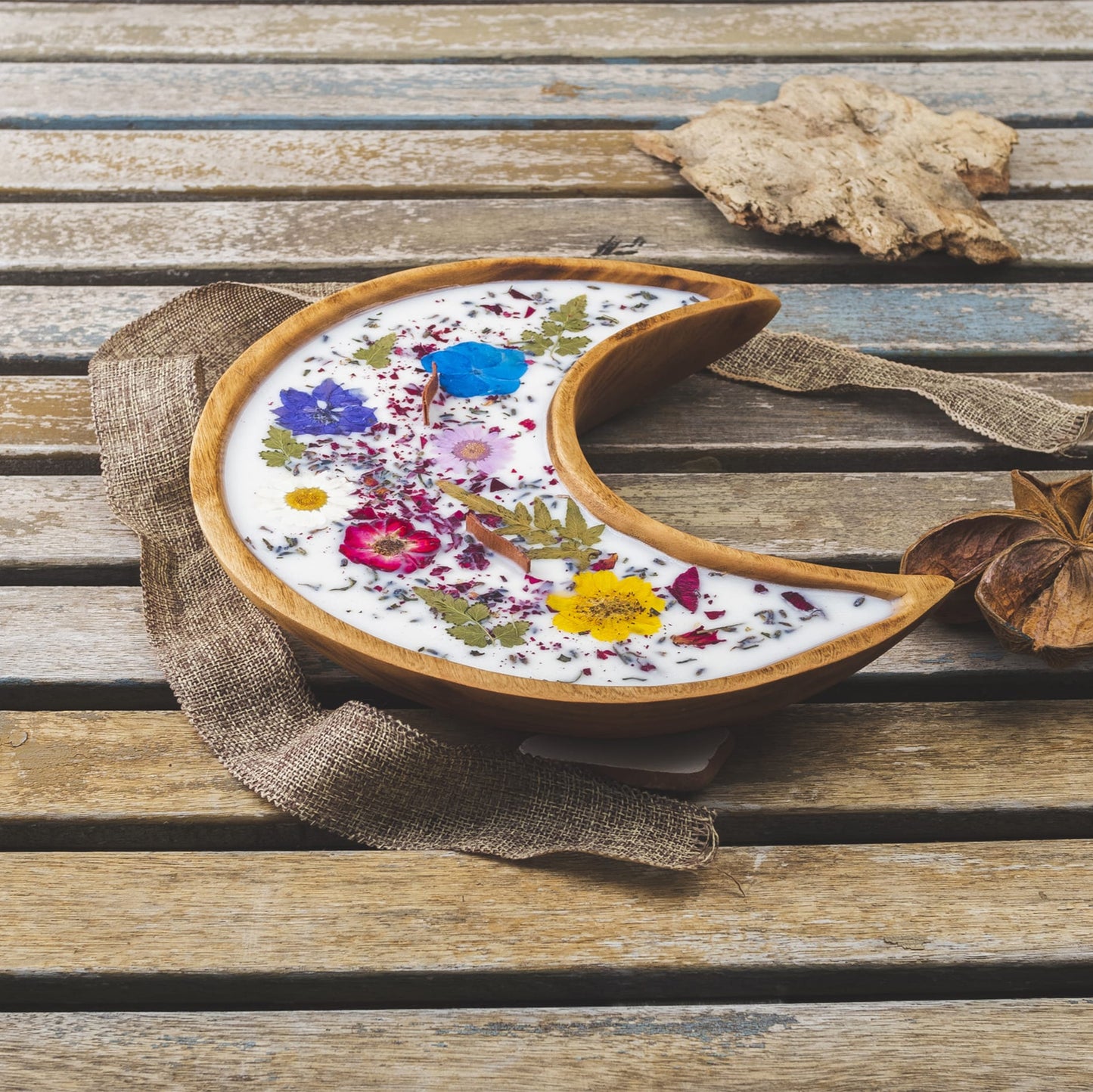 La Vela Aromática Fleur-de-Bois en Cuenco de Madera en Forma de LUNA con Mecha Ondulada, decorada con flores y hojas secas, reposa sobre madera rústica junto a una cinta de arpillera y ramas secas.