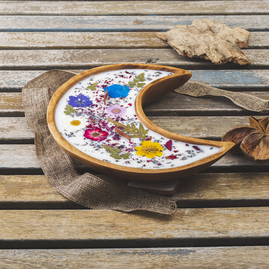 La Vela Aromática Fleur-de-Bois en Cuenco de Madera en Forma de LUNA con Mecha Ondulada, decorada con flores y hojas secas, reposa sobre madera rústica junto a una cinta de arpillera y ramas secas.