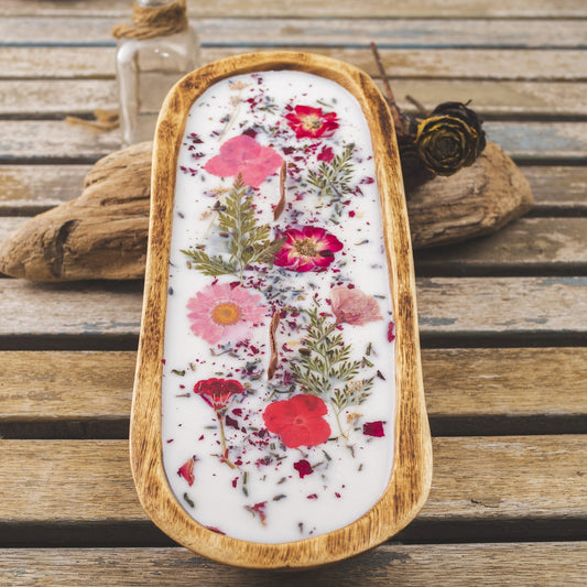 La Vela Aromatica en Cuenco de Madera Grande con Mecha Ondulada de Fleur-de-Bois, hecha de cera blanca y decorada con flores secas rojas y rosas, resalta sobre una superficie rústica junto a un frasco pequeño y una flor seca.