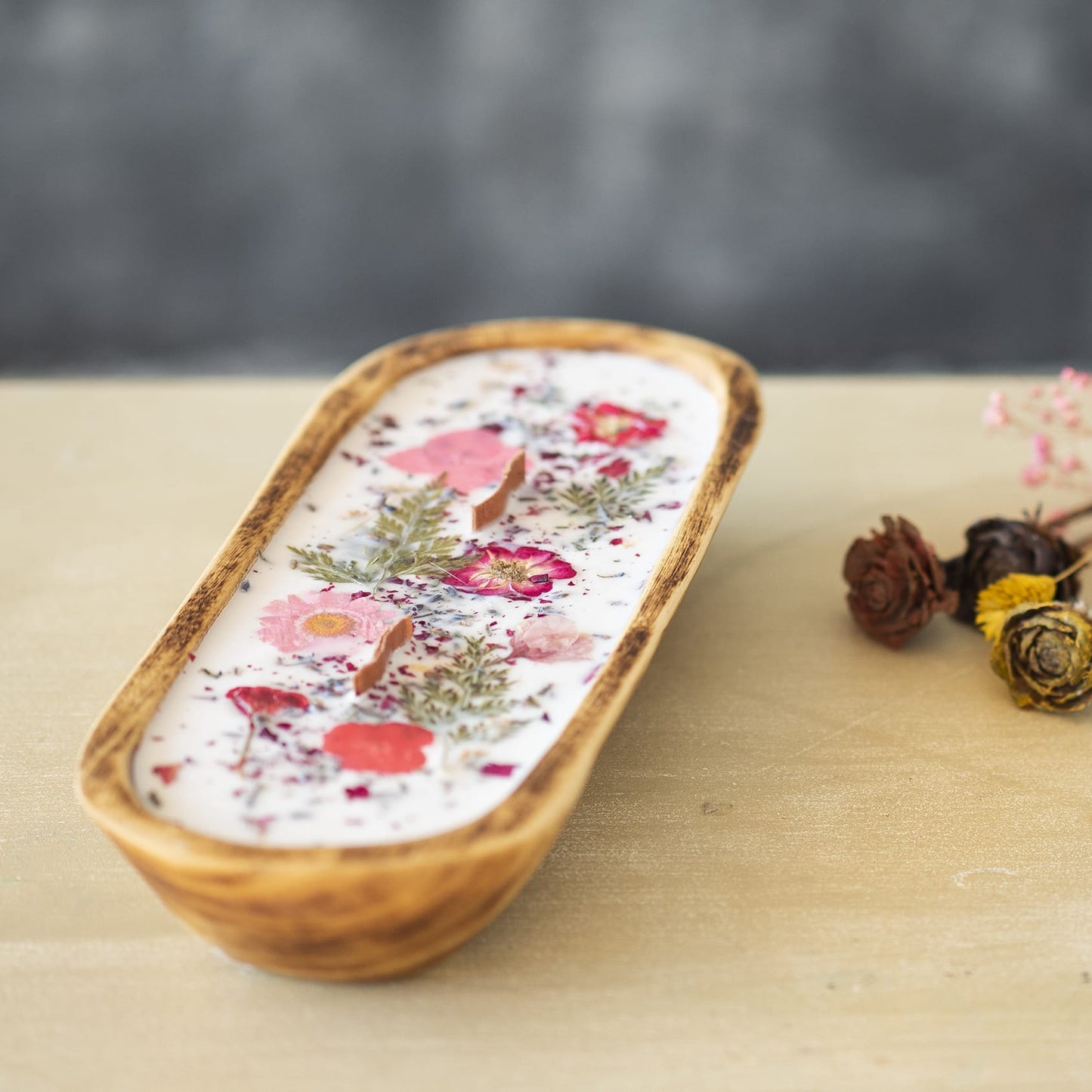 La Vela Aromatica en Cuenco de Madera Grande con Mecha Ondulada de Fleur-de-Bois es una vela perfumada de cera de soja en cuenco ovalado, decorada con flores y hierbas secas, ideal para ambientes elegantes y naturales.