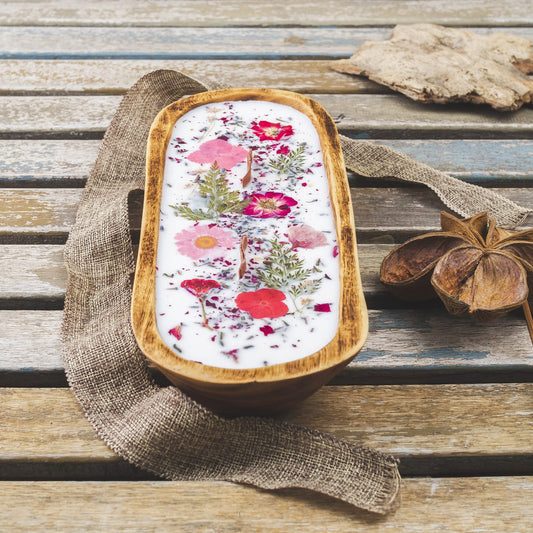 La Vela Aromatica en Cuenco de Madera Grande con Mecha Ondulada de Fleur-de-Bois, decorada con flores secas rosadas y hojas verdes, descansa sobre una mesa rústica junto a tela marrón texturizada y anís estrellado.