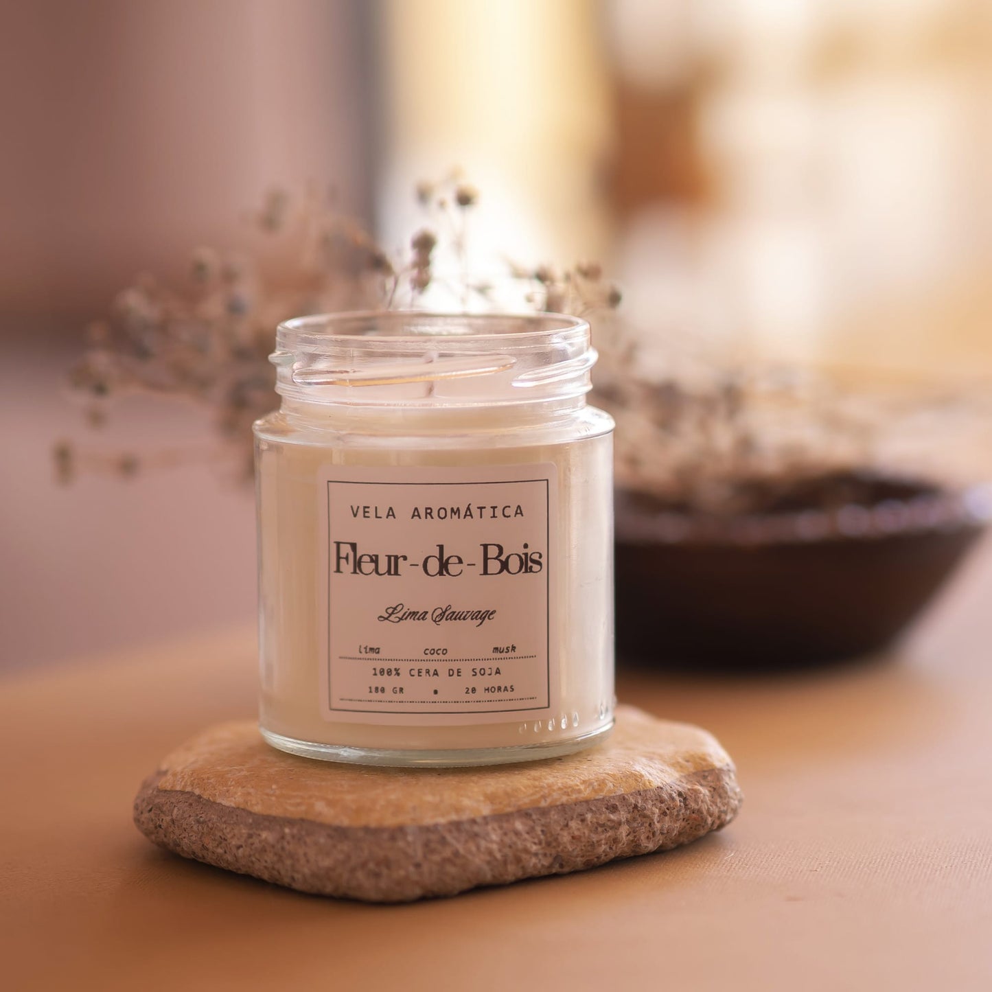 La Vela Aromática Fleur-de-Bois Signature - 180g de Fleur-de-Bois está colocada sobre una losa de piedra con flores secas y un cuenco marrón difuminado al fondo, añadiendo un toque cálido y acogedor a la decoración del hogar.