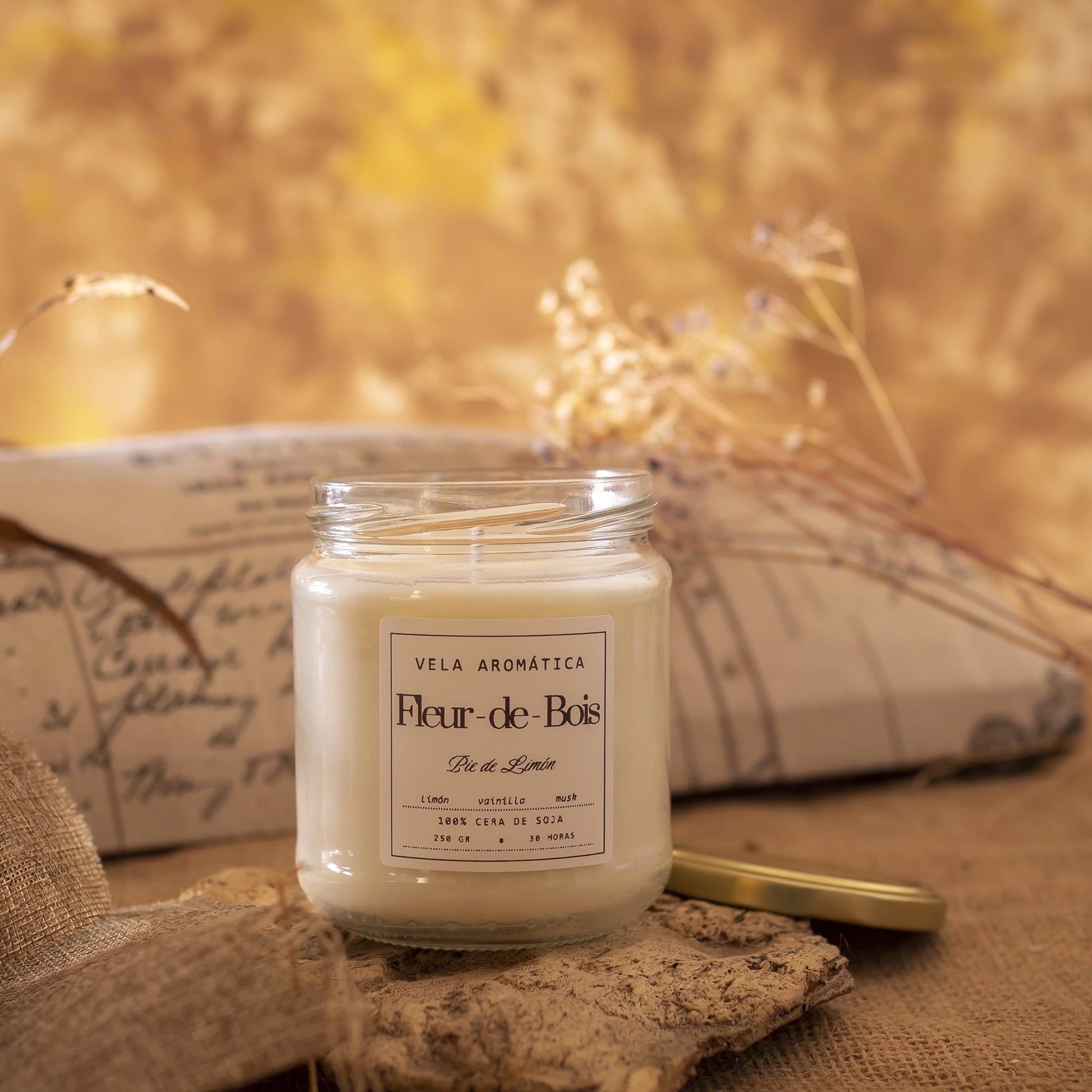 La Vela Aromática Fleur-de-Bois Signature (250 g) está colocada sobre arpillera con su tapa dorada al lado. Las plantas secas y el suave fondo marrón amarillento realzan el ambiente cálido y rústico.