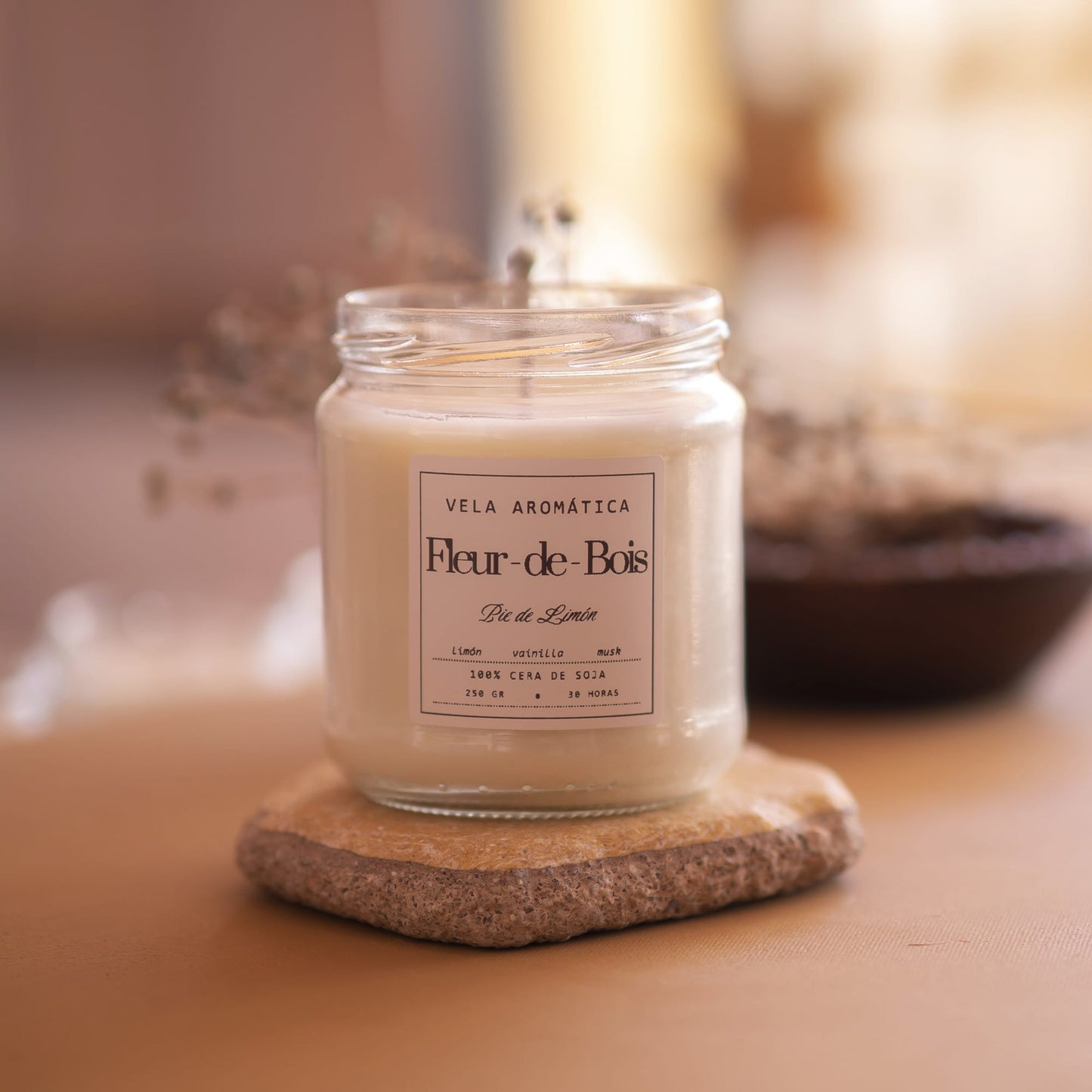 La Vela Aromática Firma Fleur-de-Bois - 250g, una vela de soja de color blanco cremoso en un tarro de cristal, descansa sobre un posavasos de piedra junto a flores secas. La elegante vela de Fleur-de-Bois destaca sobre el fondo suavemente difuminado.