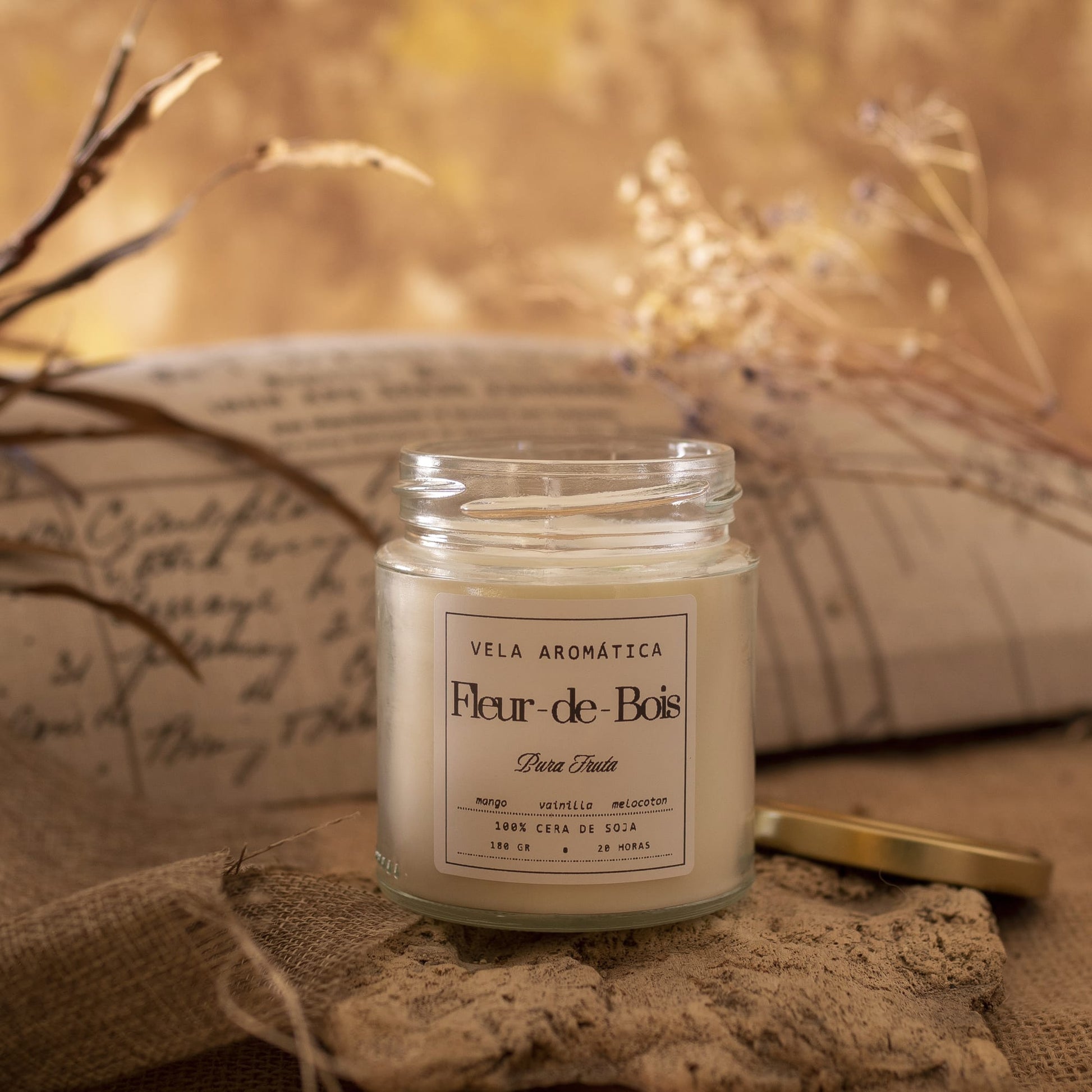 Una Vela Aromática Fleur-de-Bois - 180g se encuentra abierta sobre una superficie rústica con plantas secas y una almohada al fondo, su tapa dorada cerca, creando una cálida y acogedora decoración hogar.
