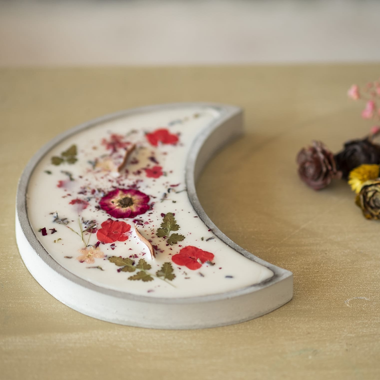 La Vela Aromática Fleur-de-Bois en Cuenco de Cemento en Forma de LUNA con Mecha Ondulada descansa sobre madera clara junto a flores secas, ideal como regalo especial o para quienes buscan una vela artesanal única.