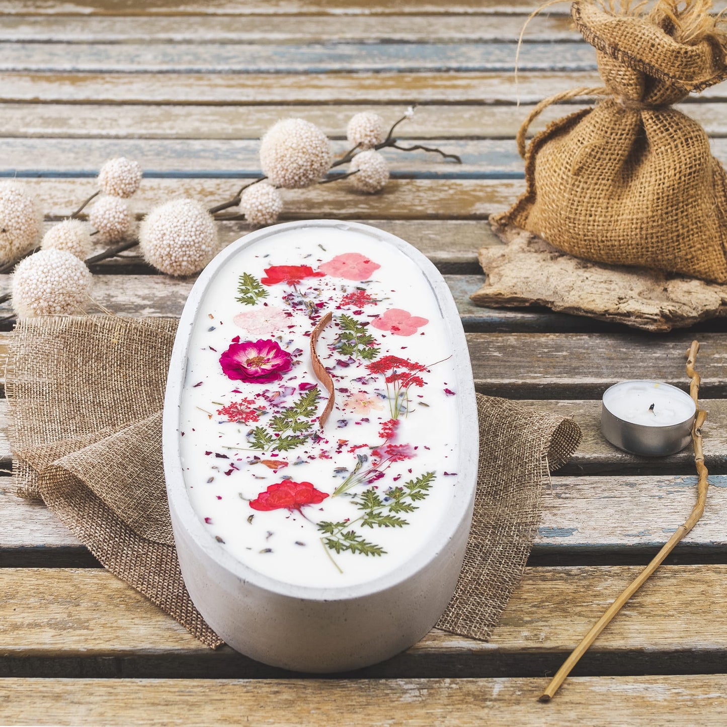 La Vela Aromática en Cuenco de Cemento Ovalado GRANDE con Mecha Ondulada de Fleur-de-Bois, decorada con flores y hojas secas, reposa sobre arpillera en una mesa rústica junto a un saco, ramas con pompones blancos y una vela tealight cercana.