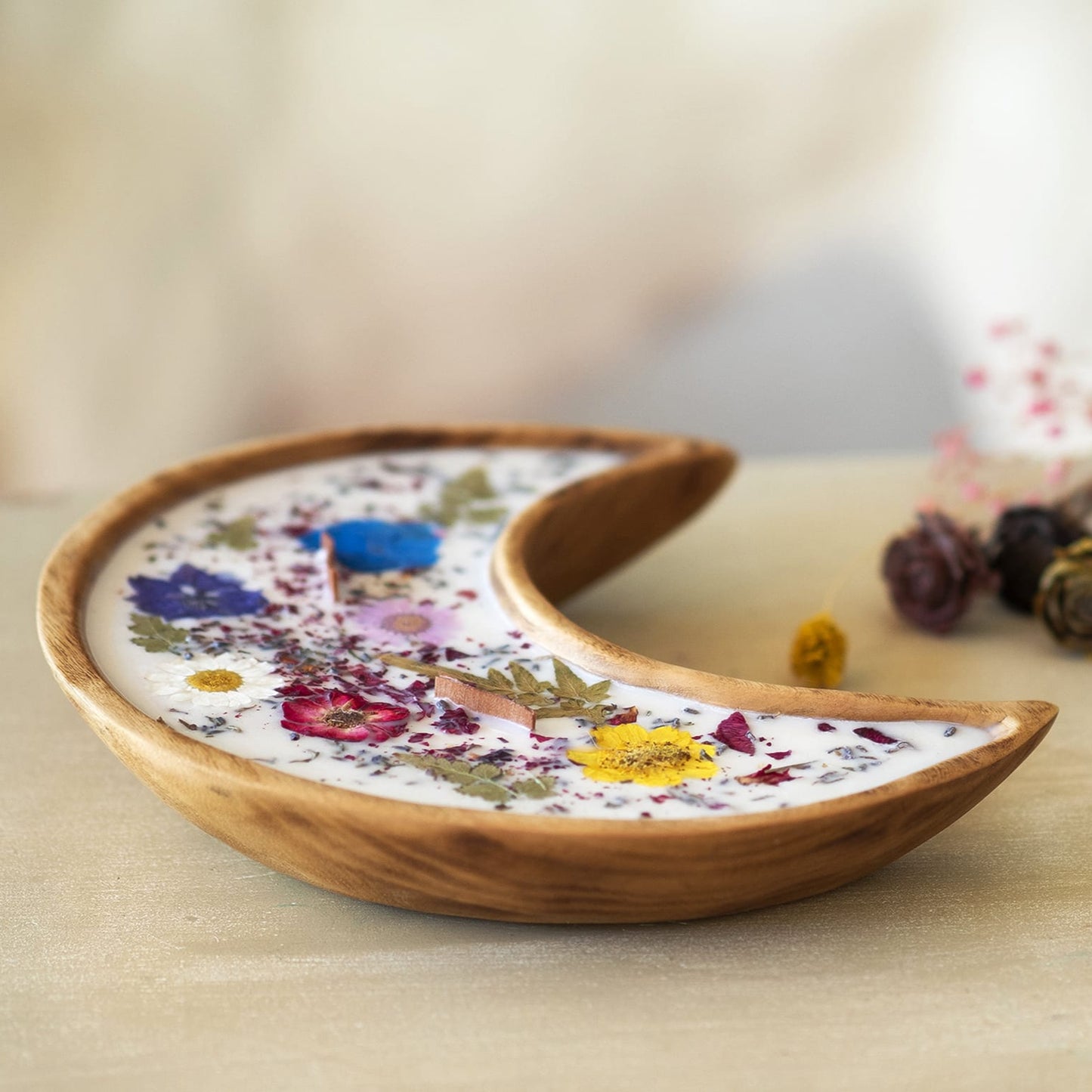La Vela Aromática Fleur-de-Bois en Cuenco de Madera en Forma de LUNA con Mecha Ondulada, hecha con cera de soja blanca y decorada con flores y hojas secas coloreadas, reposa sobre una superficie clara con flores secas desenfocadas al fondo.