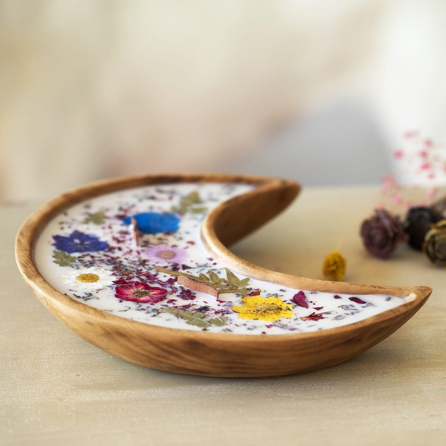 La Vela Aromática Fleur-de-Bois en Cuenco de Madera en Forma de LUNA con Mecha Ondulada, hecha con cera de soja blanca y decorada con flores y hojas secas coloreadas, reposa sobre una superficie clara con flores secas desenfocadas al fondo.