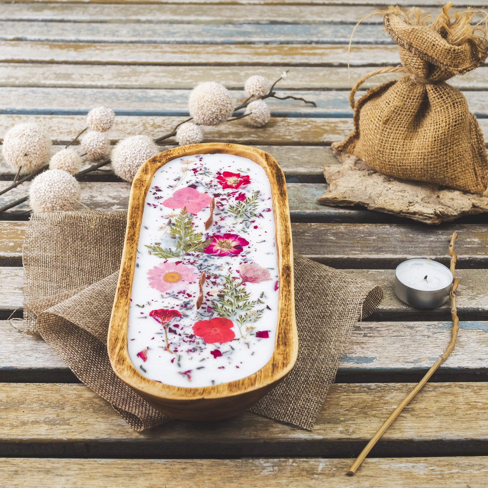 A Fleur-de-Bois Vela Aromatica en Cuenco de Madera Grande con Mecha Ondulada, decorada con flores y hierbas secas, adorna una mesa rústica junto a bolsitas de yute, ramas decorativas, una vela tealight y tela marrón.