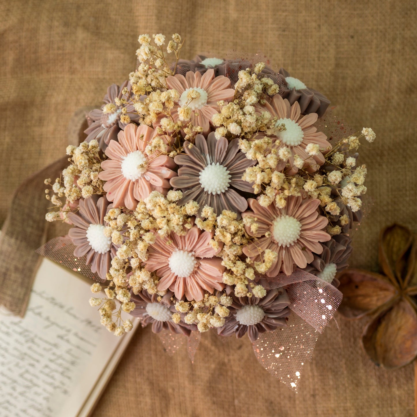 El Ramo de Flores Margaritas Clásico de Cera de Soja DOLCE CAFFÉ de Fleur-de-Bois, con flores en forma de margarita y aliento de bebé seco, se encuentra cerca de velas de soja florales sobre tela marrón texturizada junto a una carta manuscrita y una vaina de semillas.