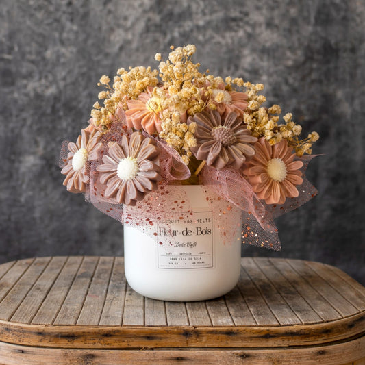 Un tarro Fleur-de-Bois blanco con la etiqueta "Ramo de Flores Margaritas Clásico de Cera de Soja DOLCE CAFFÉ" contiene flores de cera de soja de color rosa pastel, melocotón y beige con aliento de bebé amarillo seco sobre una superficie de madera rústica y un fondo gris.