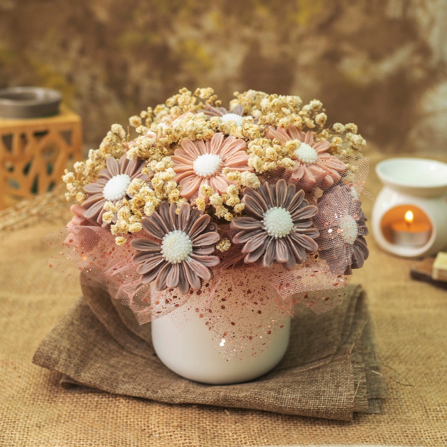 Un jarrón blanco contiene el Ramo de Flores Margaritas Clásico de Cera de Soja Fleur-de-Bois DOLCE CAFFÉ de Fleur-de-Bois en rosa, marrón y beige con centros blancos, colocado sobre arpillera con velas florales de cera y decoración rústica.