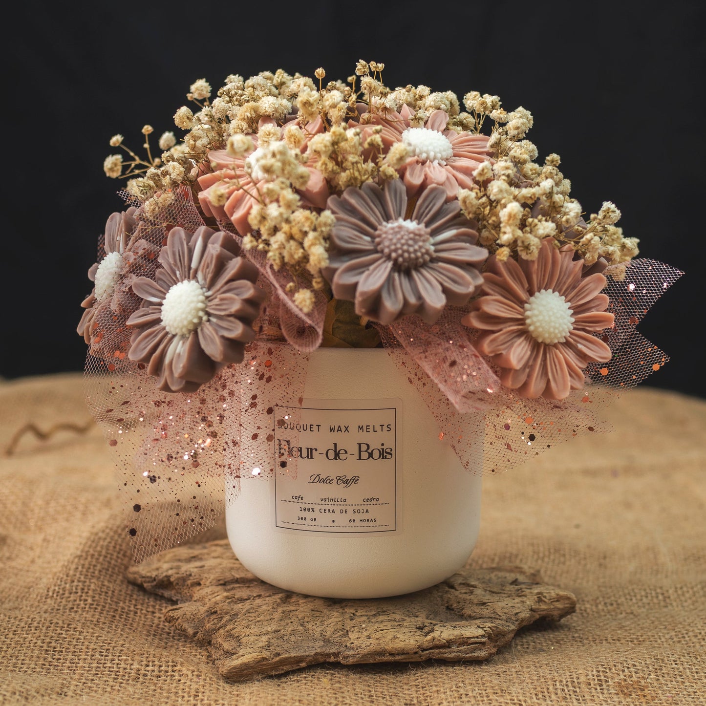 Un tarro blanco con la etiqueta Fleur-de-Bois contiene el Ramo de Flores Margaritas Clásico de Cera de Soja DOLCE CAFFÉ, adornado con tul rosa, purpurina y aliento de bebé seco para un regalo romántico, sobre un fondo oscuro.