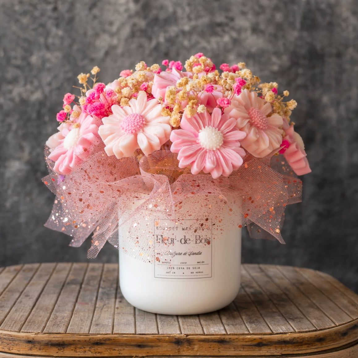 El Ramo de Flores Fleur-de-Bois Margaritas Clásico de Cera de Soja DULZURA DE SANDÍA presenta flores artificiales rosas y blancas con cinta y acentos secos, dispuestas cerca de velas derretidas sobre madera para una exhibición romántica.