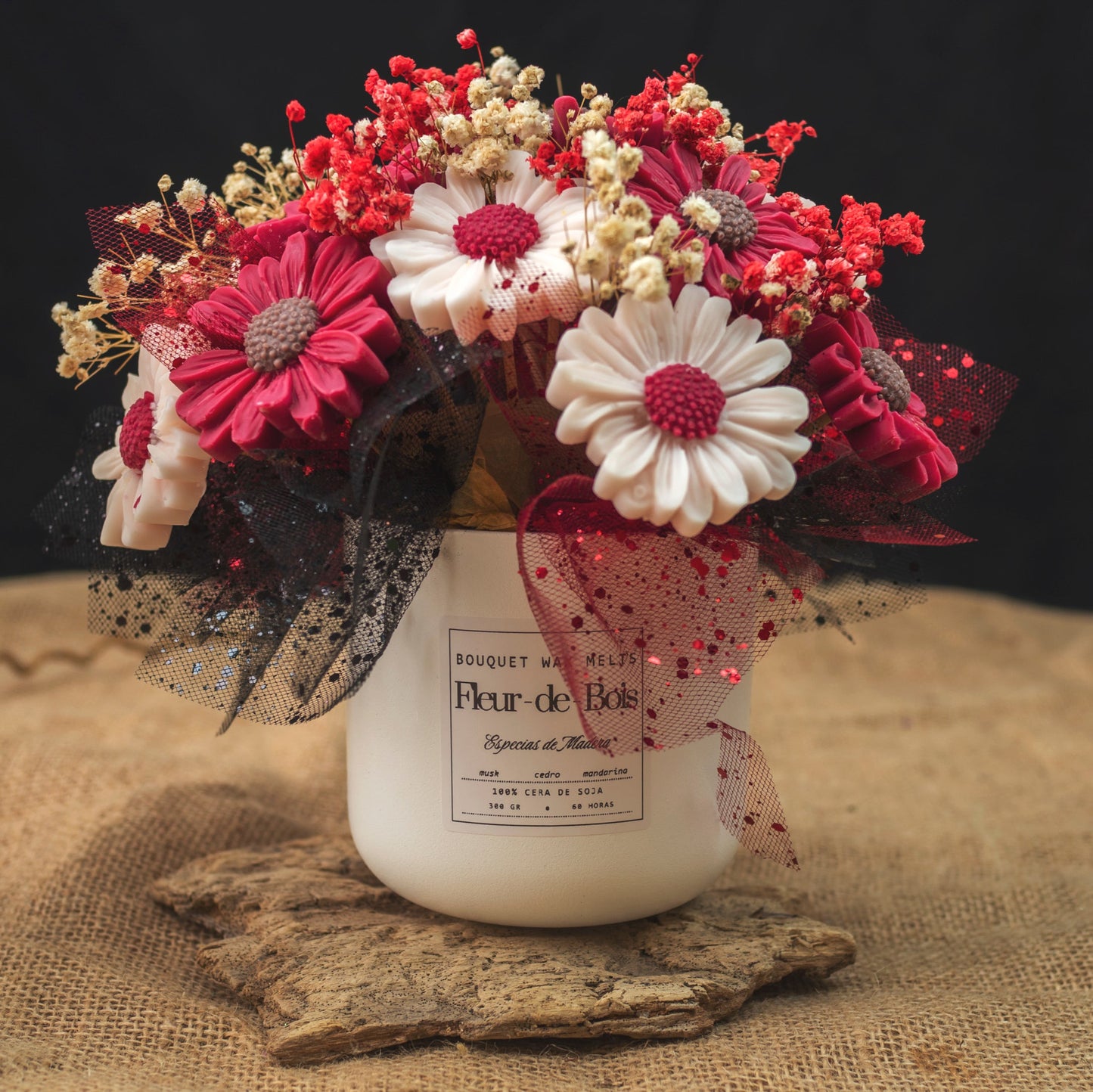 Un tarro blanco de Flor de Bois contiene el Ramo de Flores Margaritas Clásico de Cera de Soja ESPECIAS DE MADERA, con flores rojas, blancas y rosas con malla negra y aliento de bebé, colocadas sobre arpillera sobre un fondo oscuro.