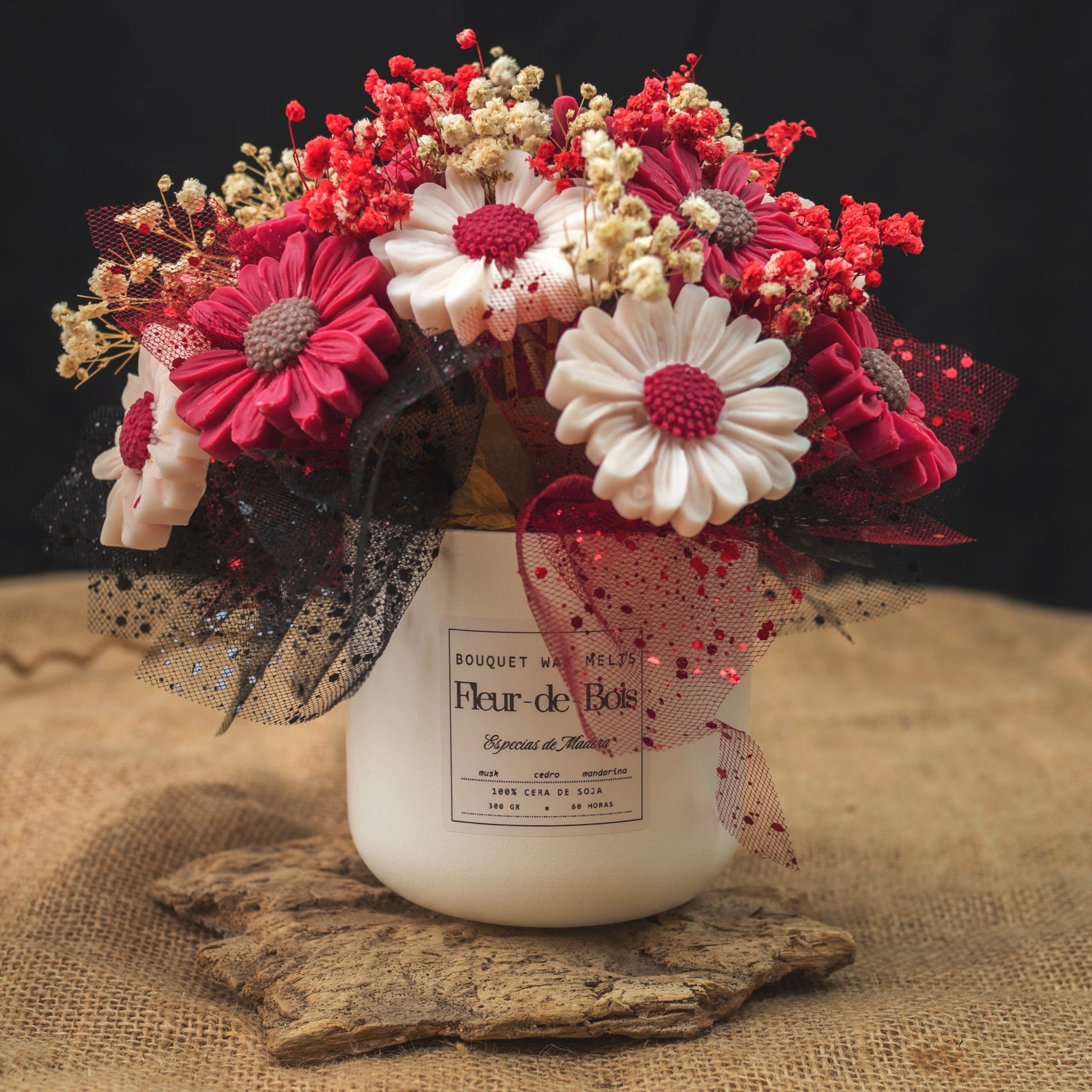 Un tarro blanco de Flor de Bois contiene el Ramo de Flores Margaritas Clásico de Cera de Soja ESPECIAS DE MADERA, con flores rojas, blancas y rosas con malla negra y aliento de bebé, colocadas sobre arpillera sobre un fondo oscuro.