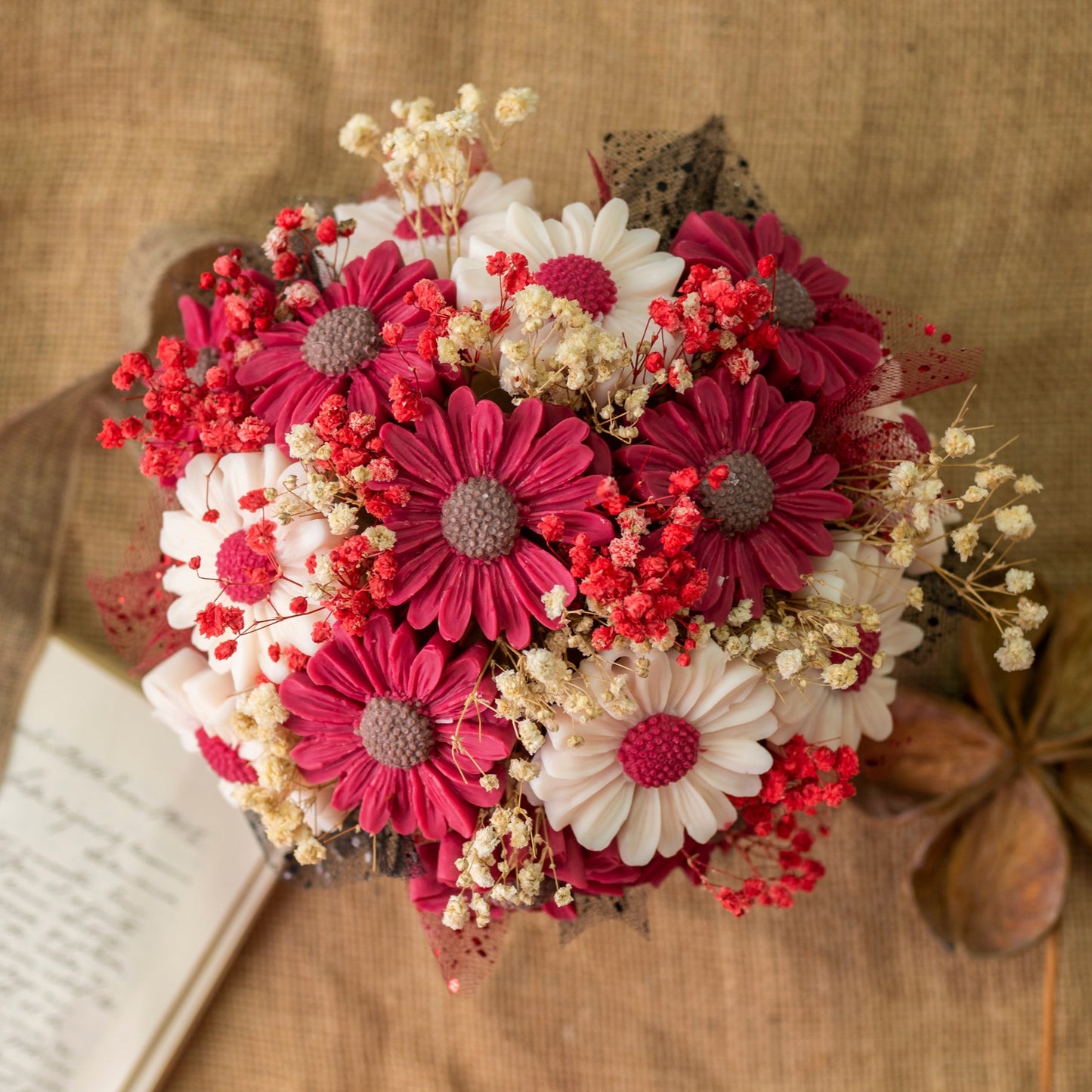 El Ramo de Flores Margaritas Clásico de Cera de Soja Fleur-de-Bois de Fleur-de-Bois, con margaritas y flores secas, está colocado sobre arpillera junto a velas florales, un libro abierto y una cinta marrón, ideal como regalo romántico.