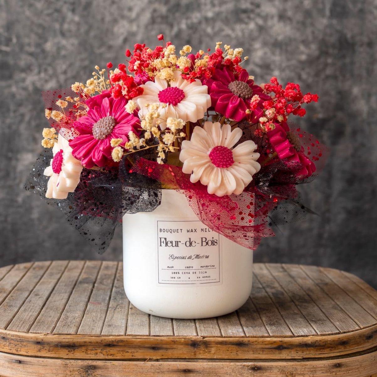 Un tarro blanco Flor de Bois contiene el Ramo de Flores Margaritas Clásico de Cera de Soja ESPECIAS DE MADERA -flores artificiales rojas, rosas y blancas- sobre madera rústica con fondo gris texturizado. Un regalo romántico ideal.