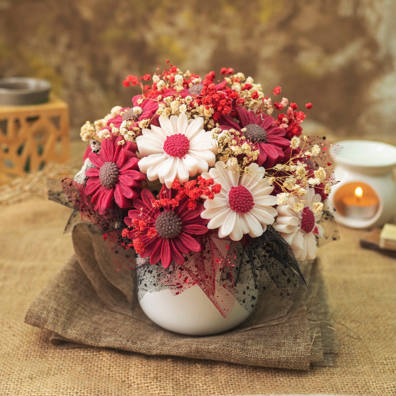 Un jarrón blanco sostiene el Ramo de Flores Fleur-de-Bois Margaritas Clásico de Cera de Soja ESPECIAS DE MADERA en rosa, blanco y rojo con acentos secos y malla negra, colocado sobre arpillera con velas brillantes, es un perfecto regalo romántico.