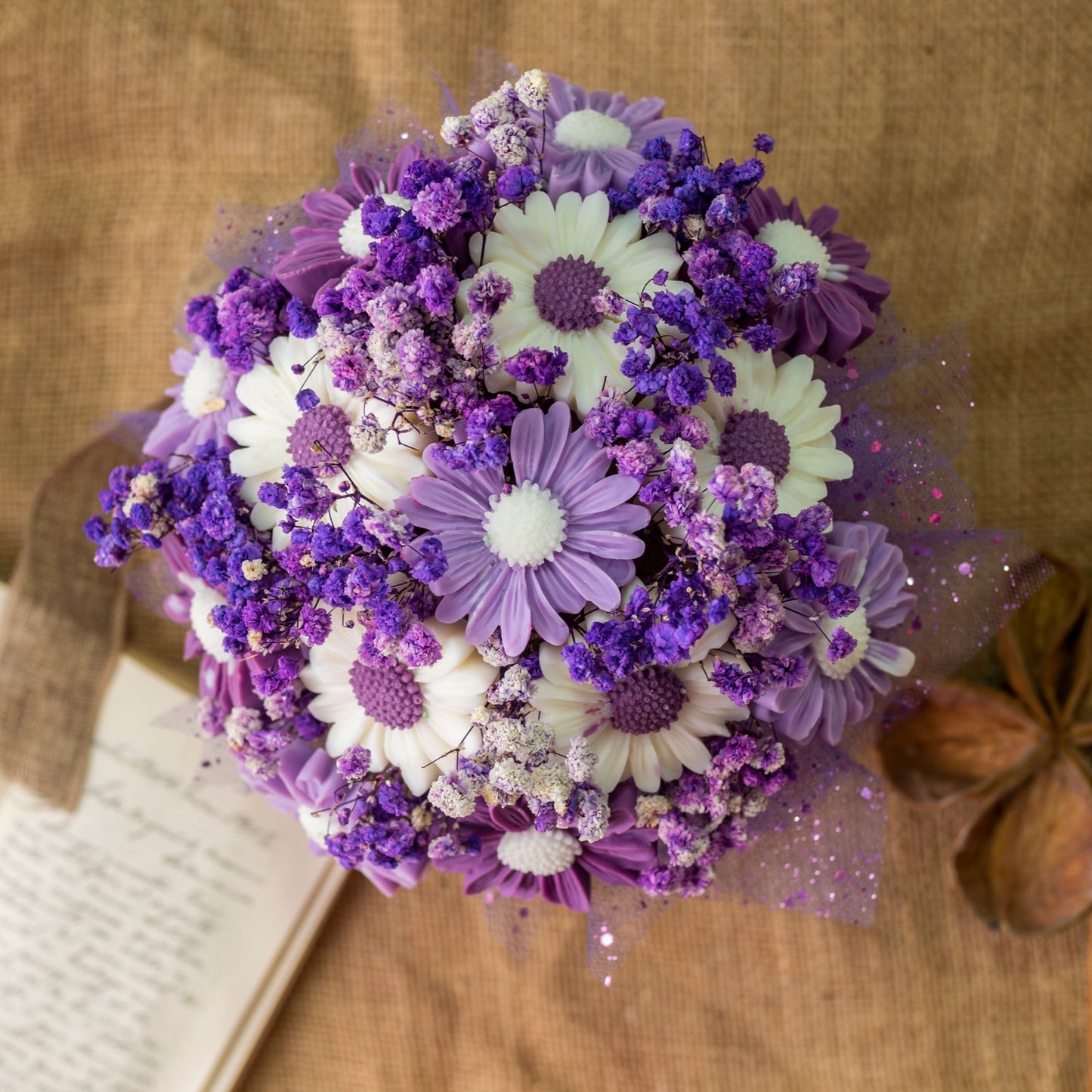 El Ramo de Flores Margaritas Clásico de Cera de Soja FRESCURA DE LAVANDA de Fleur-de-Bois, con margaritas blancas y moradas y pequeñas flores moradas, descansa sobre arpillera junto a un cuaderno abierto, constituyendo un encantador regalo romántico.