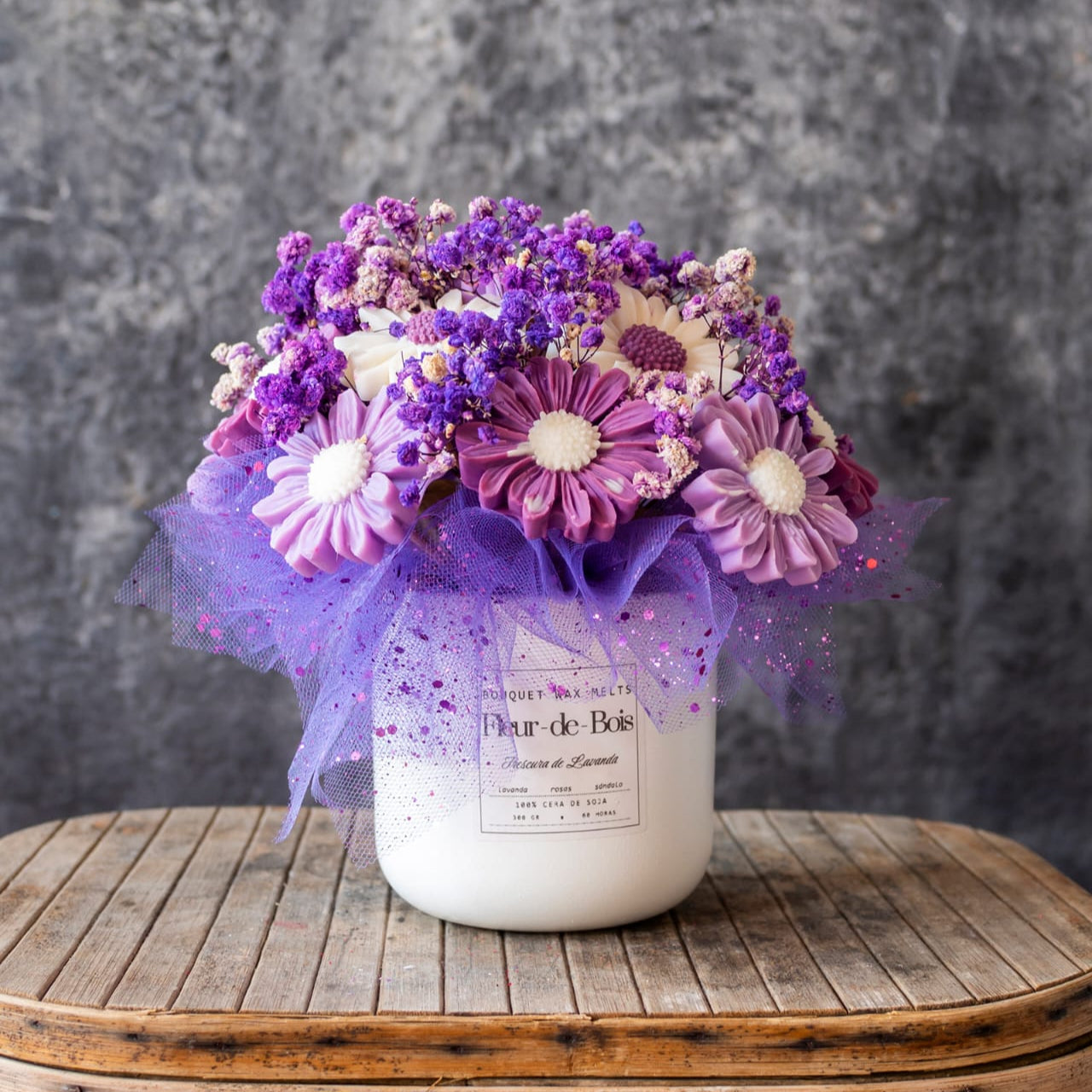 Un jarrón blanco Flor de Bois sostiene un Ramo de Flores Margaritas Clásico de Cera de Soja FRESCURA DE LAVANDA, acentuado con tul morado sobre una superficie de madera y fondo gris-un regalo romántico ideal.
