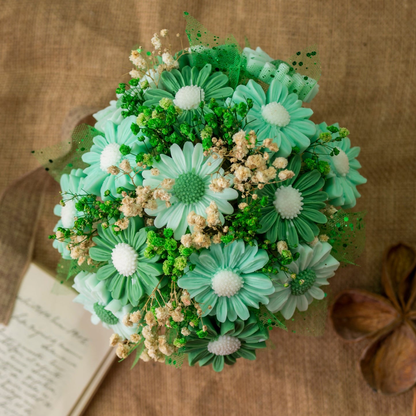 El Ramo de Flores Margaritas Clásico de Cera de Soja Fleur-de-Bois LIMA SAUVAGE de Fleur-de-Bois presenta margaritas artificiales de color verde azulado y blanco, pequeñas flores blancas, follaje verde y flores de cera de soja dispuestas sobre arpillera junto a un cuaderno.