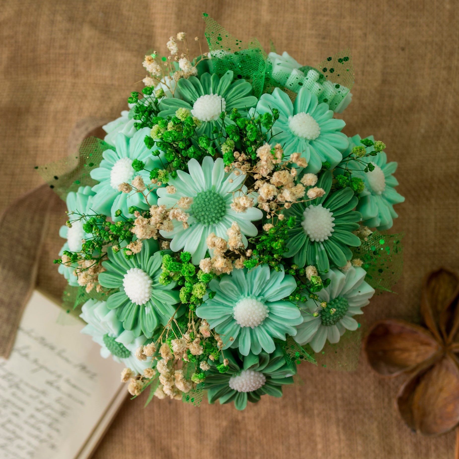 El Ramo de Flores Margaritas Clásico de Cera de Soja Fleur-de-Bois LIMA SAUVAGE de Fleur-de-Bois presenta margaritas artificiales de color verde azulado y blanco, pequeñas flores blancas, follaje verde y flores de cera de soja dispuestas sobre arpillera junto a un cuaderno.