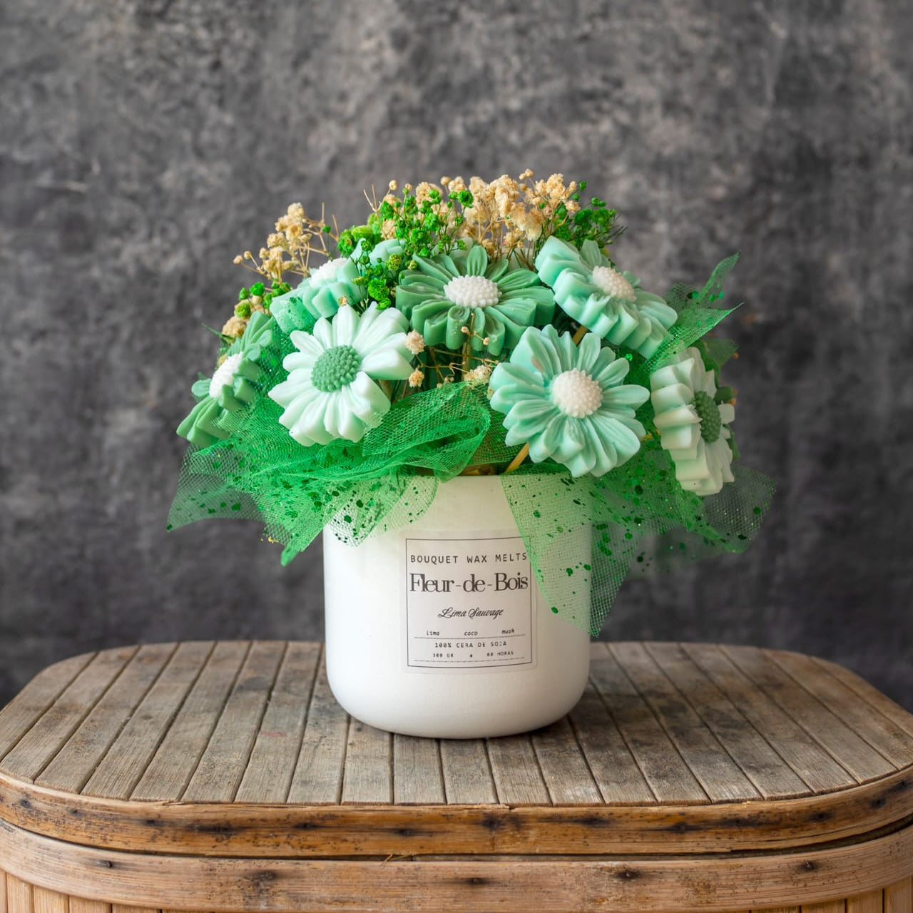 Una maceta blanca Fleur-de-Bois sostiene el Ramo de Flores Margaritas Clásico de Cera de Soja Fleur-de-Bois LIMA SAUVAGE, con flores artificiales verdes y blancas, con detalles de malla verde, sobre una mesa de madera con fondo gris.