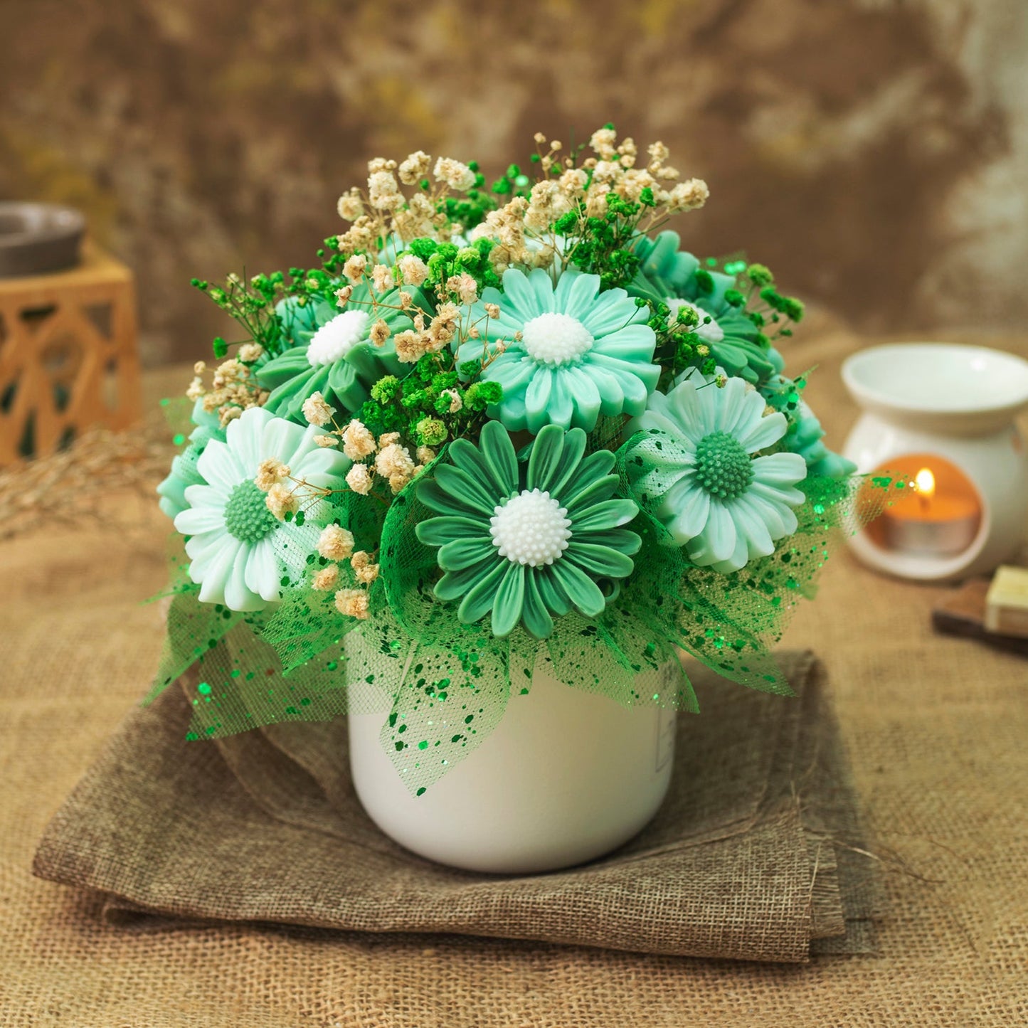 La Flor de Flores Ramo de Flores Margaritas Clásico de Cera de Soja LIMA SAUVAGE, con margaritas verde azulado y blancas, rellenos crema, acentos verdes y flores de cera de soja en un jarrón blanco sobre arpillera. Una vela encendida y la decoración se difuminan suavemente en el fondo.